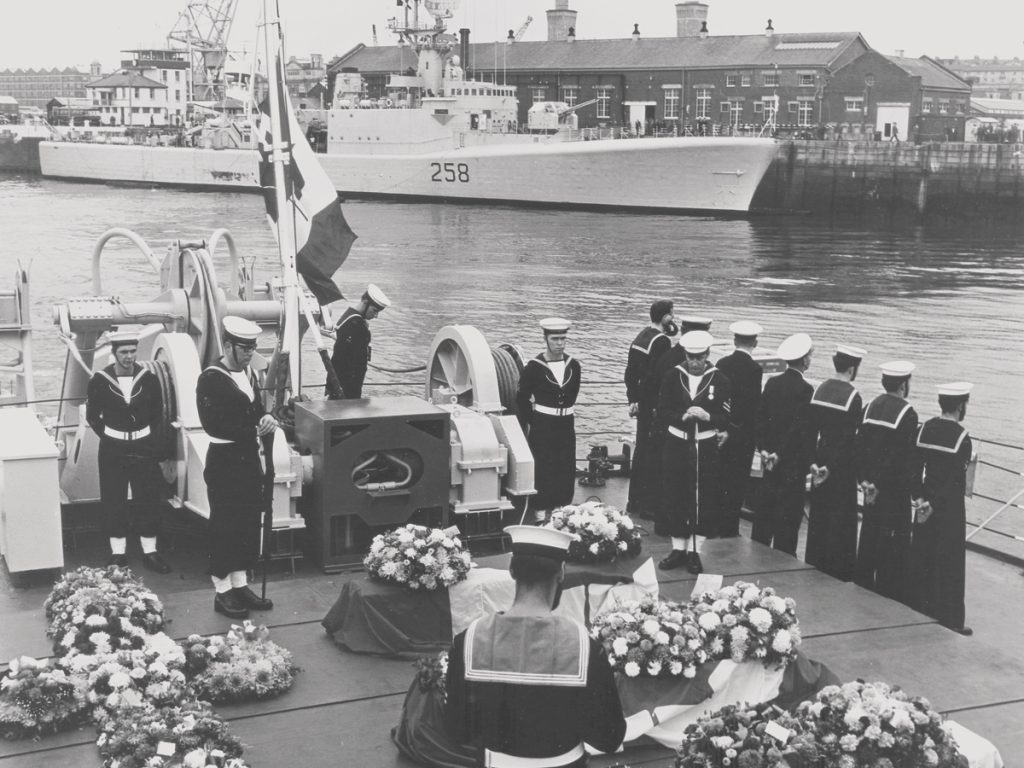 Funeral service for the nine deceased crew members of HMCS Kootenay with the burnt and damaged ship in the background at Devonport